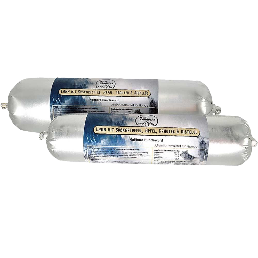 Zwei haltbare Hundewürste in silberner Kunststoffhülle. Das Etikett trägt die Aufschrift „Lamm mit Süßkartoffel, Apfel, Kräuter & Distelöl“ auf dunkelblauem Hintergrund mit Bergmotiv.