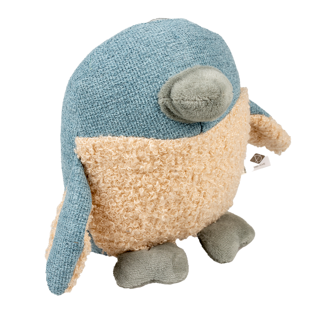 Plüschspielzeug in Vogeloptik mit blauem Stoff an Kopf, Flügeln und Rücken; beige, lockige Struktur am Bauch; grauer Schnabel und Füße, kleines Etikett am Flügel.