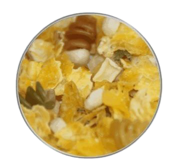 Eine gelbliche Flockenmischung mit vereinzelten weißen Würfeln und kleinen, spiralförmigen Nudeln in Grün und Braun, gleichmäßig verteilt in einer kreisrunden Ansicht.