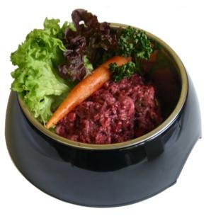 Ein schwarzer Napf mit gewolftem Lamm-Mix in dunkelrot-brauner Farbe und faseriger Struktur, ergänzt durch eine dekorative Karotte und etwas Blattgrün.