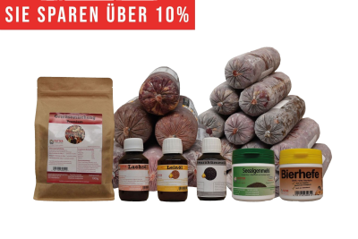 Ein vielseitiges BARF-Paket mit pyramidenförmig gestapelten Frostfleischrollen in grauem Kunstdarm. Davor stehen Flachbeutel mit rotem Inhalt, ein brauner Standbeutel, vier kleine Fläschchen und zwei Dosen.