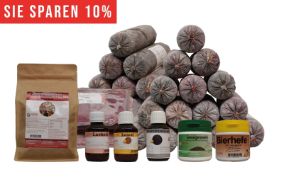 Ein vielseitiges BARF-Paket mit pyramidenförmig gestapelten Frostfleischrollen in grauem Kunstdarm. Davor stehen Flachbeutel mit rotem Inhalt, ein brauner Standbeutel, vier kleine Fläschchen und zwei Dosen.