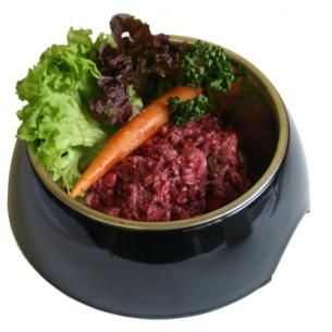 Lamm-Mix Supreme 7 x 1000g Ein schwarzer Napf mit gewolftem Lamm-Mix in dunkelrot-brauner Farbe und faseriger Struktur, ergänzt durch eine dekorative Karotte und etwas Blattgrün.